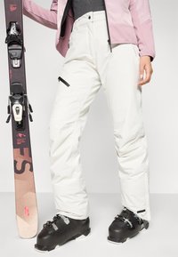 Hvit sn�øbukse med rett snitt, glidelåslommer og svarte detaljer, kombinert med svarte skistøvler og en ski. Stoffet ser ut til å være værbestandig.