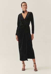 Črna žametna obleka z dolgimi rokavi, do sredine teleta, z pasom za samostojno vezanje. Ustrezajo ji črne pete z ankle trakovi in drzne uhane.