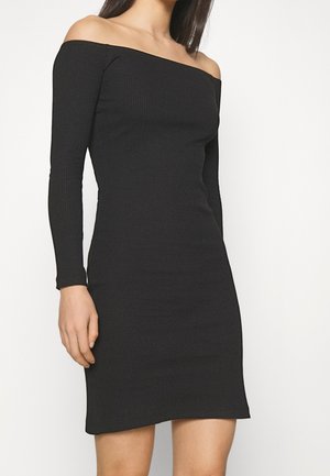 Vrouw die een zwarte, geribbelde, off-shoulder, nauwsluitende jurk met lange mouwen draagt, staand tegen een effen witte achtergrond.