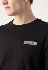 Melna kokvilnas t-kreklu ar apaļu kakla izgriezumu, kurā ir sudraba taisnstūra uzlīme ar melnu tekstu "Calvin Klein". Mīksta tekstūra, vienkāršs dizains.
