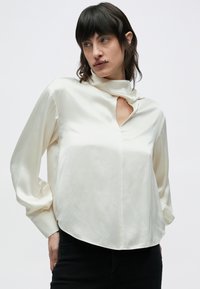 Kremowa satynowa bluzka z dekoltem typu keyhole, długimi rękawami i zaokrąglonym dołem. Charakteryzuje się gładką teksturą i luźnym krojem.