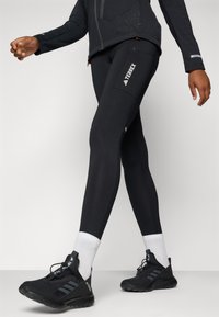 Leggings de sport noirs à coupe ajustée, dotés d'une poche latérale et du logo "Terrex" en blanc. Associés à des chaussures légères noires et des chaussettes blanches.