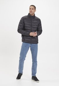 Veste noire matelassée avec un col haut, présentant une fermeture éclair avant et un design matelassé horizontal, associée à un jean bleu clair et des baskets noires.