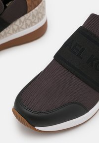 Chaussure de sport noire et marron avec une tige en tissu texturé, une sangle élastique avec logo, une semelle extérieure en caoutchouc et un détail à motif au talon.