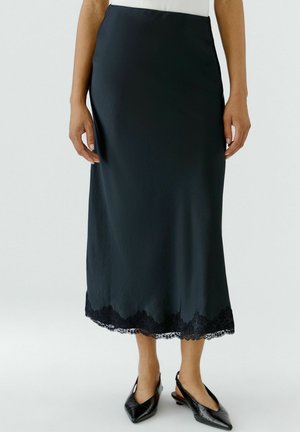Jupe midi noire avec bordure en dentelle portée par une personne debout, associée à des chaussures slingback noires à bout pointu sur un fond clair.