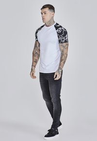 Bílý tričko s černými vzorovanými krátkými rukávy, spárované s tmavými slim-fit džíny a černými teniskami. Na rukou jsou viditelné tetování.