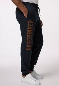 Navy sweatpants i mjukt material, med en brun "BASERUNNER" tryck längs sidan, och utrustade med fickor och en justerbar midja.