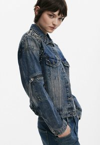 Džínová bunda v tmavomodrej farbe so striebornými cvočkami na pleciach, predných vreckách a detailmi na zips na rukávoch. S ošúchaným povrchom a klasickým golierom.