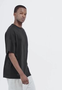 Young man in black t-shirt and light gray pants walking, facing left, on plain white background.