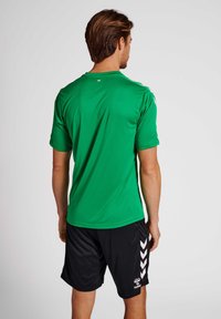Grünes sportliches Trikot mit kurzen Ärmeln; hat einen Rundhalsausschnitt und eine glatte Textur. Kombiniert mit schwarzen Shorts, die ein weißes Zickzackmuster zeigen.