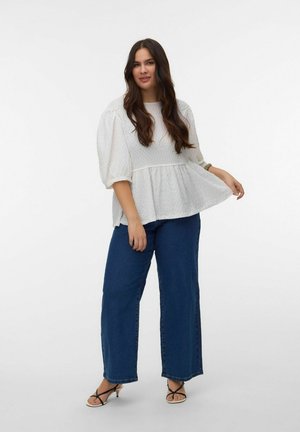 Kobieta o długich ciemnych włosach nosi białą, teksturowaną bluzkę z bufkami, niebieskie szerokie jeansy i czarne sandały na obcasie na jednolitym tle.