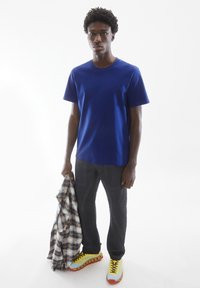 Blaues Baumwoll-T-Shirt, kurze Ärmel, kombiniert mit dunklen Hosen und mehrfarbigen Turnschuhen. Er hält ein gemustertes Flanellhemd.