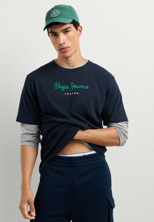 Junger Mann, der eine grüne Kappe und ein dunkelblaues Pepe Jeans London T-Shirt trägt, zieht es hoch, um graue Langarmshirts und dunkelblaue Hosen mit einer Tasche zu zeigen.