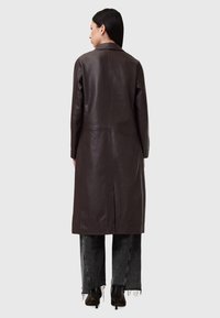 Cappotto in pelle marrone con taglio dritto, maniche lunghe e colletto a revers, abbinato a jeans neri sfilacciati e tacchi.