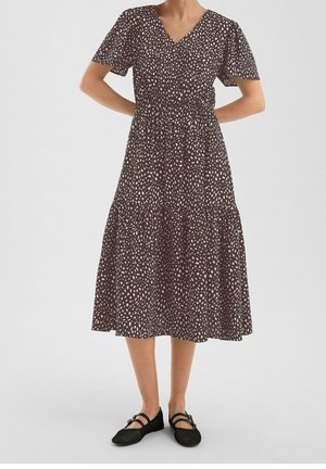 Kvinde iført en midi sort kjole med hvide prikker, korte ærmer og sorte Mary Jane flade sko, stående med hænderne bag ryggen.