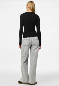 Žena s dugom plavom kosom, koja nosi usku crnu majicu dugih rukava, svijetlosive široke traperice i cipele sa smešnim đonom, okrenuta je leđima.