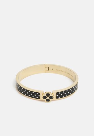 Bransoletka typu bangle w złotym odcieniu z czarną emalią i białymi kropkami, z zapięciem w kształcie czterolistnej koniczyny w kolorze czarnym.