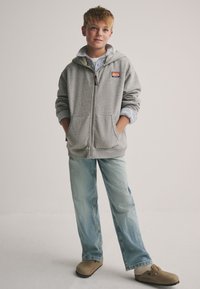 Grauer Kapuzenpullover mit Reißverschluss und einer Vordertasche, darüber ein blau-weiß gestreiftes Hemd. Hellblaue Jeans und beige Clogs runden das Outfit ab.