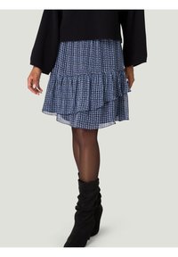 Jupe bleue à motifs avec des volants superposés, en tissu léger. Portée avec des bottes noires molles jusqu'aux genoux et un haut noir texturé.