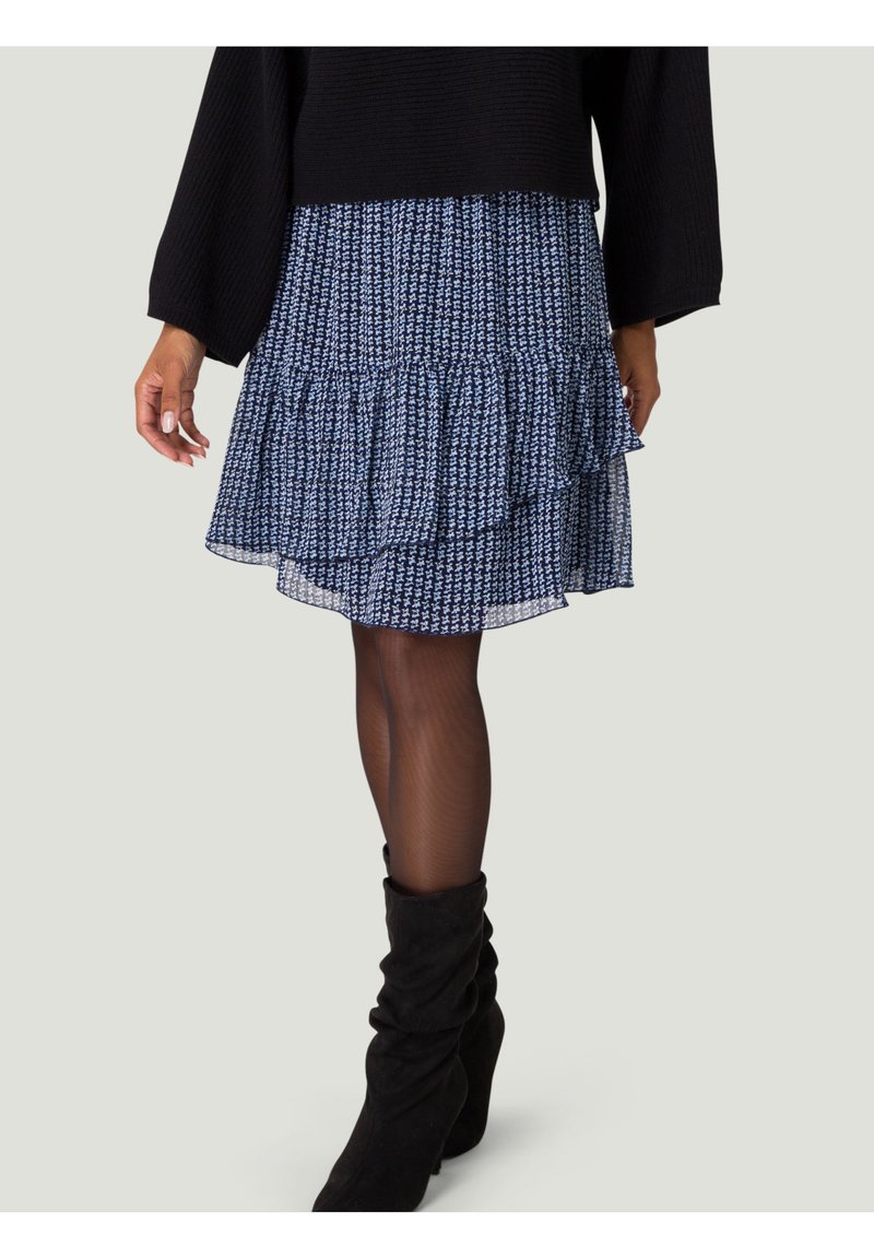 Jupe bleue à motifs avec des volants superposés, en tissu léger. Portée avec des bottes noires molles jusqu'aux genoux et un haut noir texturé.