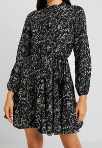 Svart blommig klänning med hög krage, långa ärmar, elastisk midja och lager på lager- kjol med vita blommiga mönster.