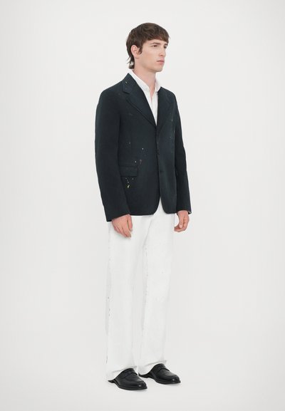 Blazer negro con manchas de pintura, camisa blanca, pantalones blancos y zapatos negros. Modelo de pie contra un fondo blanco liso.