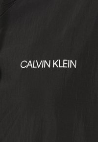 Svart jacka med textilstruktur och minimalistisk design; inkluderar vit "CALVIN KLEIN"-logotyp framtill.