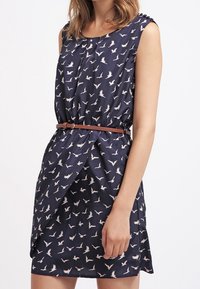 Laivastonsininen mekko, jossa on valkoisia lintukuvioita, olkaimeton malli, kiristetty vyötäisiltä ruskealla vyöllä ja kietaisumallinen etuosa. Silkkinen kangastuntu.