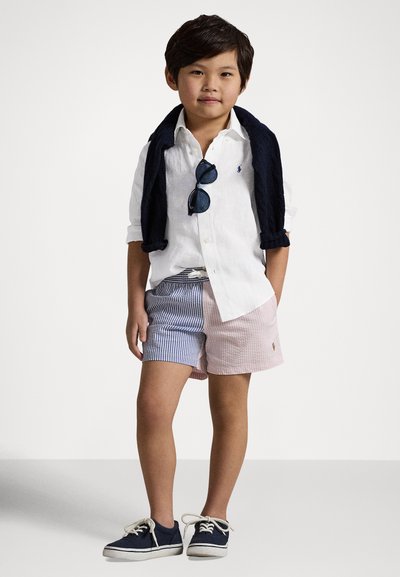 Λευκό πουκάμισο με κοντά μανίκια, πάνω ένα μπλε πουλόβερ ριγμένο στους ώμους, ριγέ σορτς σε μπλε και ροζ αποχρώσεις, μπλε αθλητικά παπούτσια, μαύρα γυαλιά ηλίου, στέκεται μπροστά σε ένα απλό φόντο.