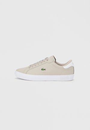 Beige synthetic sneaker with round toe, white rubber sole, and lace-up design. Features green Lacoste logo on side and contrasting heel accent.