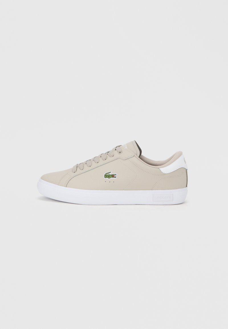 Beige syntetisk sneaker med rund tå, hvid gummisål og snørelukning. Har et grønt Lacoste-logo på siden og en kontrasterende hælaccent.