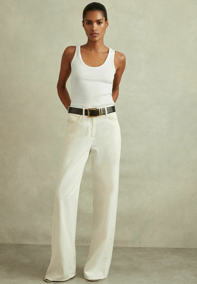 Canotta bianca abbinata a pantaloni crema a vita alta e gamba larga. Cintura nera con fibbia dorata. Tessuto dalla trama liscia, design minimale.