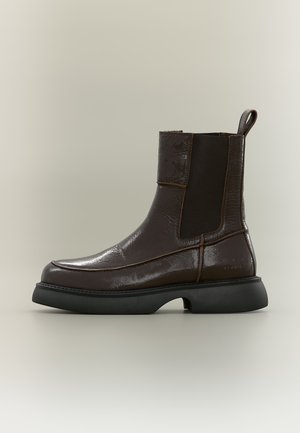 Dunkelbrauner Leder-Knöchelboot mit schwarzem elastischem Seitenpanel und klobiger schwarzer Sohle auf neutralem Hintergrund.