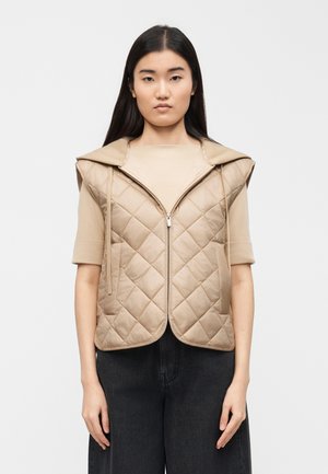 Ženska z dolgimi temnimi lasmi nosi bež prešito brezrokavnik s kapuco čez bež zgornji del s kratkimi rokavi in črne široke hlače.