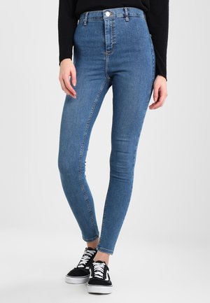 Jeans skinny de mezclilla azul de cintura alta con una textura suave, diseño de cinco bolsillos, cierre con botón frontal y combinados con zapatillas negras.