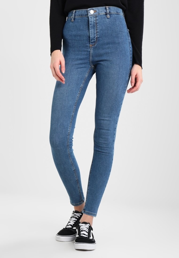 Jeans skinny de mezclilla azul de cintura alta con una textura suave, diseño de cinco bolsillos, cierre con botón frontal y combinados con zapatillas negras.