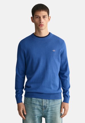 Pull en tricot bleu avec un col rond, des poignets côtelés et un ourlet côtelé. Il présente un petit logo sur la poitrine gauche. Assorti à un jean en denim bleu clair.