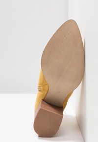 Zapato amarillo de ante con puntera afilada y tacón de bloque de madera apilado, con la suela que muestra la talla 37, posicionado verticalmente contra paredes blancas en una esquina.