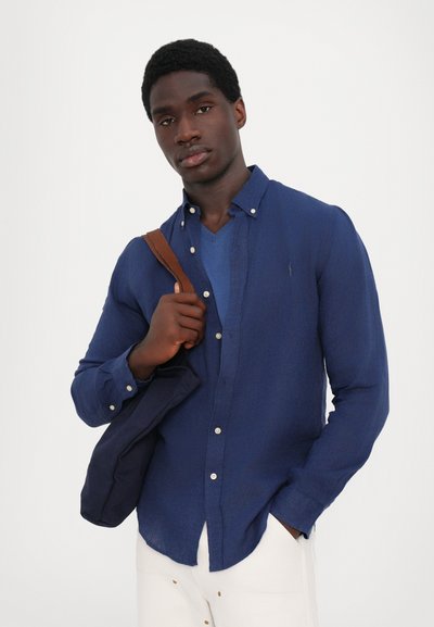 Mornarsko modra srajca z dolgimi rokavi iz teksturirane tkanine, z gumbi, kombinirana z modrim majico z V-izrezom in poudarjena s rjavim naramnim trakom.