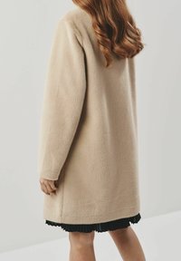 Cappotto beige in tessuto morbido con vestibilità comoda, lungo fino al ginocchio, caratterizzato da maniche lunghe e un orlo arrotondato, indossato sopra una gonna nera a pieghe.