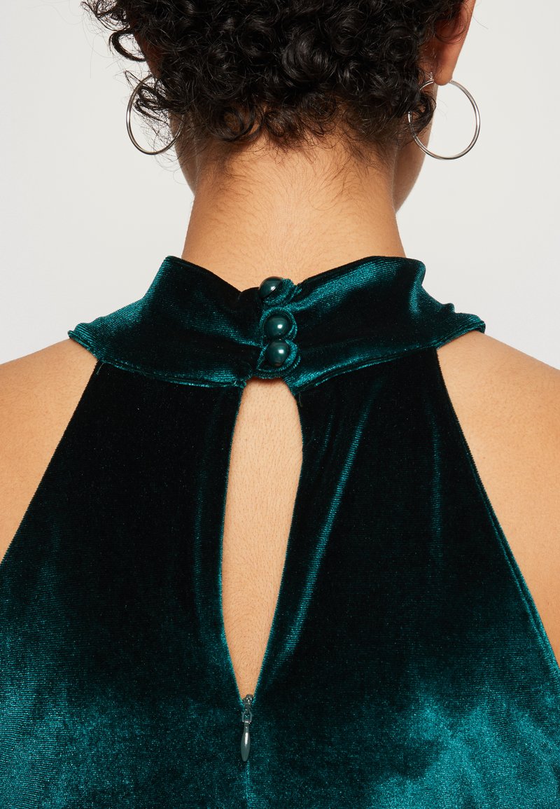 Abito in velluto verde con scollatura alta, dotato di un'apertura sul retro e tre bottoni decorativi. Texture liscia e finitura lucida.
