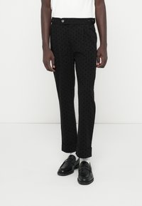 Calças pretas com um padrão de pontos, apresentando um ajuste ajustado, dois bolsos frontais e fecho com botão. Usadas com mocassins pretos.