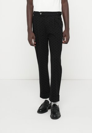 Calças pretas com um padrão de pontos, apresentando um ajuste ajustado, dois bolsos frontais e fecho com botão. Usadas com mocassins pretos.