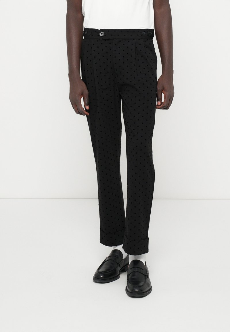 Calças pretas com um padrão de pontos, apresentando um ajuste ajustado, dois bolsos frontais e fecho com botão. Usadas com mocassins pretos.