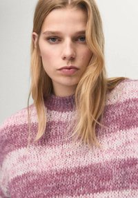 Giovane donna con lunghi capelli biondi che indossa un maglione lavorato a maglia a righe rosa e viola, che guarda dritto davanti a sé con un'espressione neutra.