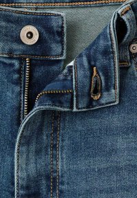 Denim-farkut, joissa on tumma pesu, kuparinväriset saumat, metallinen nappi ja vetoketjullinen läppä sekä pehmeä, hieman teksturoitu kangas.