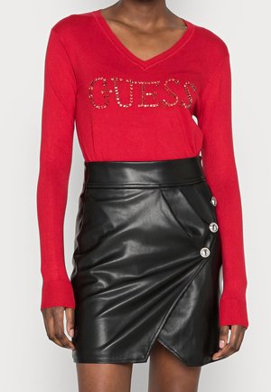 Czerwony sweter z dekoltem w serek z napisem „GUESS” zdobionym kryształkami, połączony z czarną skórzaną spódnicą z wysokim stanem, ozdobioną trzema srebrnymi guzikami po boku.