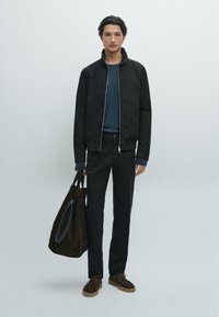 Schwarze Jacke mit hohem Kragen, blaues Hemd darunter, dunkelgraue Hose, braune Wildledertasche und passende braune Schnürschuhe.