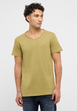 Mann mit lockigem Haar, der ein olivgrünes V-Ausschnitt-T-Shirt und dunkelblaue Jeans trägt, blickt nach links vor neutralem Hintergrund.