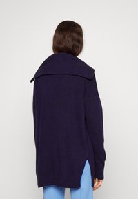 Knitted dark purple cardigan with a wide collar, long sleeves, side slits, and ribbed texture at the collar and hem. Relaxed fit.